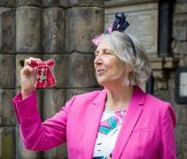 Sue Briggs, dressed in a floral dress and pink jacket, looking towards her MBE which she is holding up in front of her 