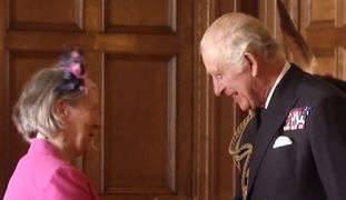 King Charles III is standing on the right and smiling down at Sue Briggs on the left.  