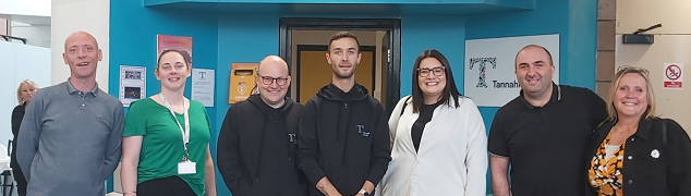 Picture of the seven people who met at Tannahill Centre, smiling and standing in a line in front of the Tannahill Centre reception area. 