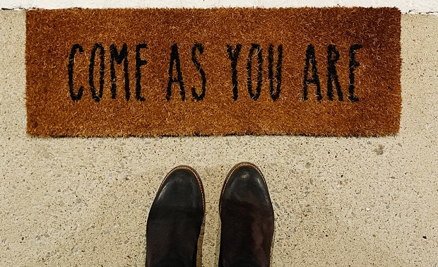 Top of the picture is a brown door mat with the words Come As You Are written in black on it. At the bottom of the picture are two black boots. 