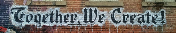 
Picture of large black graffiti on white paint, which has been done on side of a red bricked building. It says Together We Create 