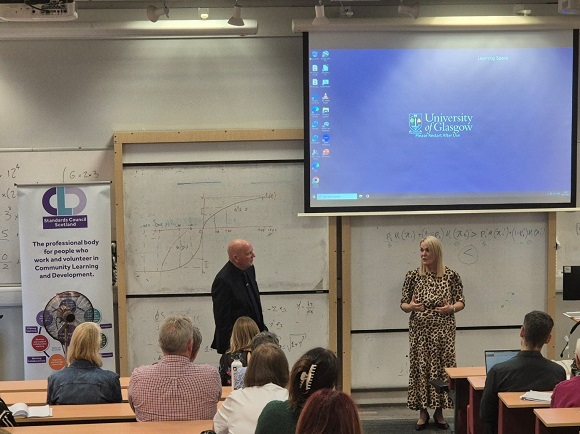 Photograph of Alan Sherry OBE and Kate Still in front  of an audience in a lecture hall with the CLD Standards Council banner and Glasgow University on presentation screen.