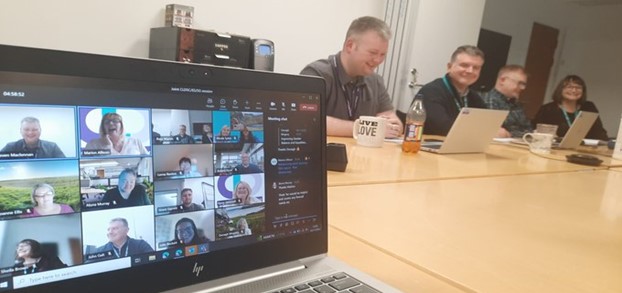 Members of the CLD Standards Council, Education Scotland HMI CLD Team sit round a desk with laptops, another laptop shows people who have joined the meeting online.