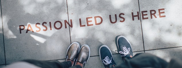 Photo looking down at two pairs of shoes, standing side by side on grey paving slabs, with the words “Passion led us here” written on the slabs.