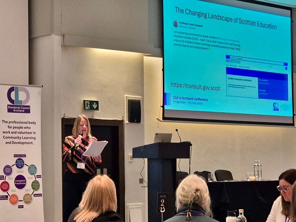 Professor Sinead Gormally holding a mic to the left of a lectern. CLD Standards Council pop up banner is behind her right shoulder. To her left, is a large screen with a page of a presentation on The Changing Landscape of Scottish Education. 