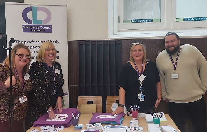 From Left to Right. Kirsty, Marion, Vikki and Chris, smiling whilst standing behind a table full of Standards Council goodies with a CLD Standards Council banner behind on the left.